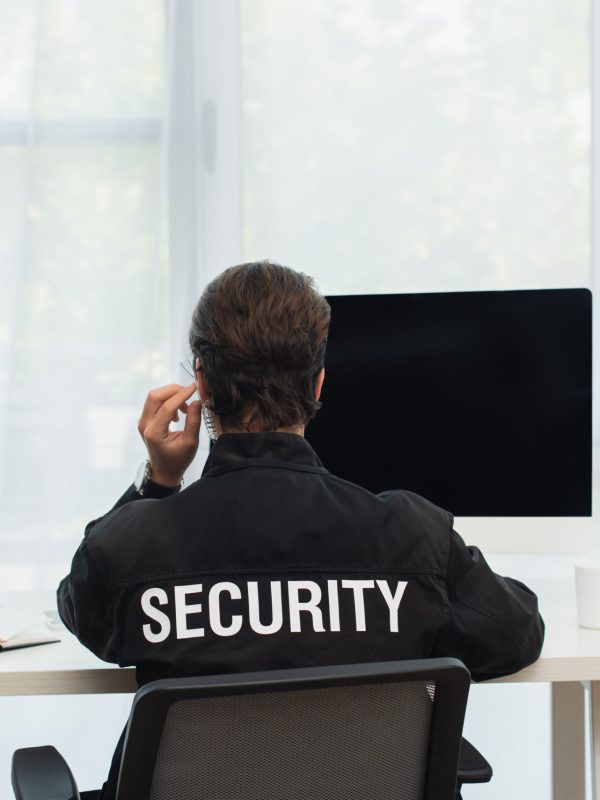 back view of security man in black uniform and earphone sitting near computers in supervision room