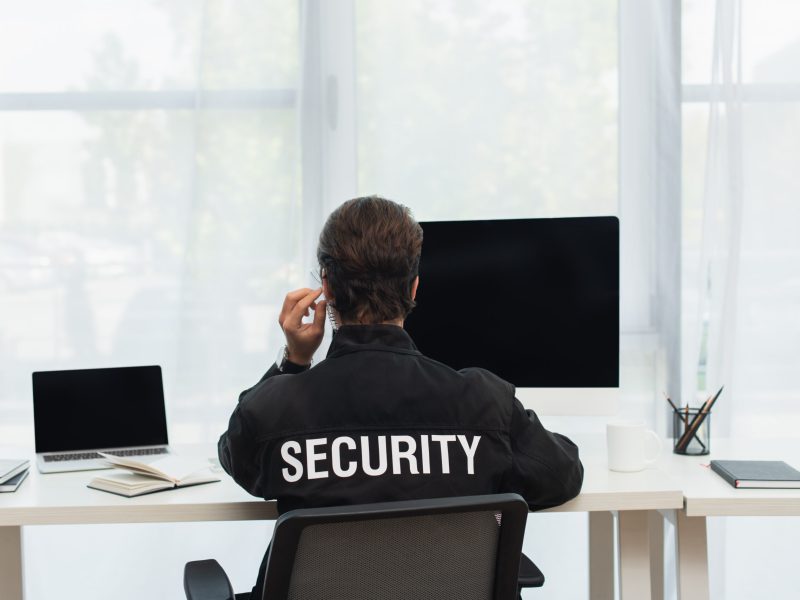back view of security man in black uniform and earphone sitting near computers in supervision room