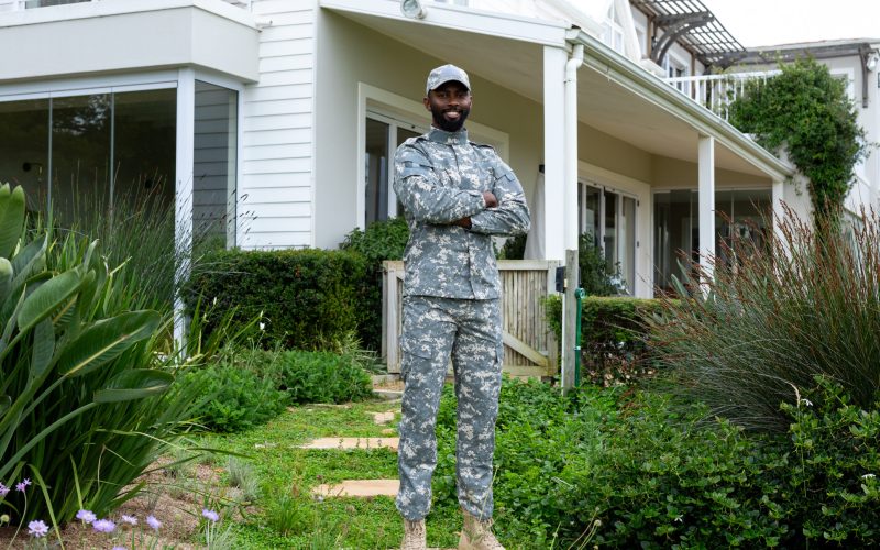 Portrait of happy african american male soldier wearing military uniform staying against house. Armed forces, lifestyle and domestic life, unaltered.