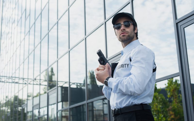 security man in sunglasses standing with weapon near building with glass facade